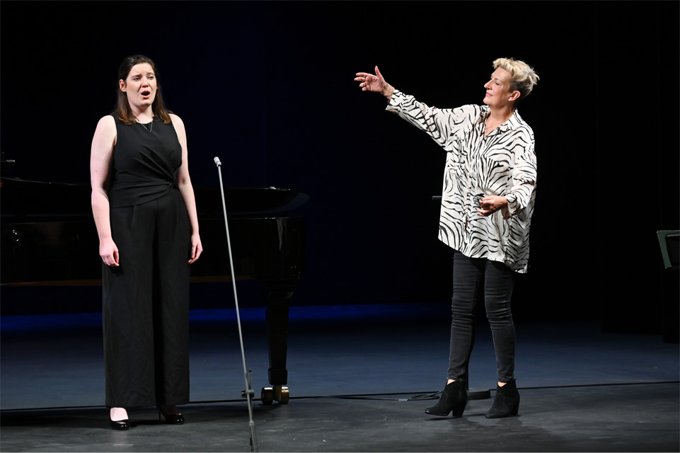 A woman with blonde hair wearing a white blouse and black trousers gesturing to a vocal student wearing all black who is singing test